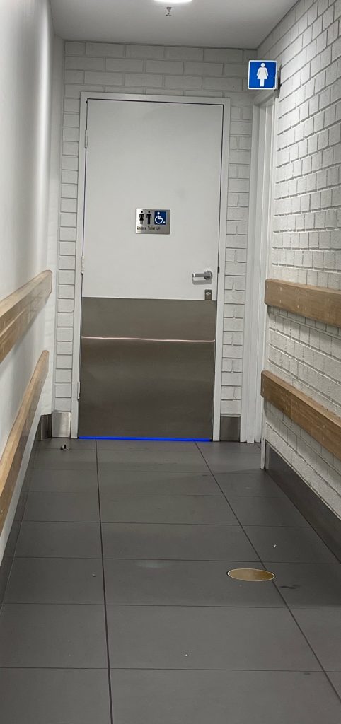 a corridor leads to the accessible toilet, blue light coming under the door, door is adjacent to the womens toilet