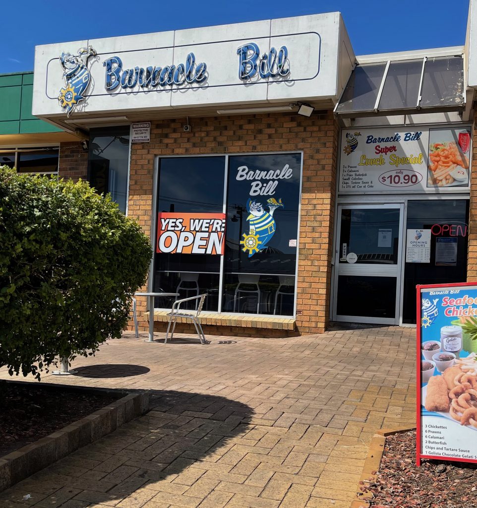 Barnacle Bills glass windows on the left, the door to the right. Pavers lead to the business and a bush to the left