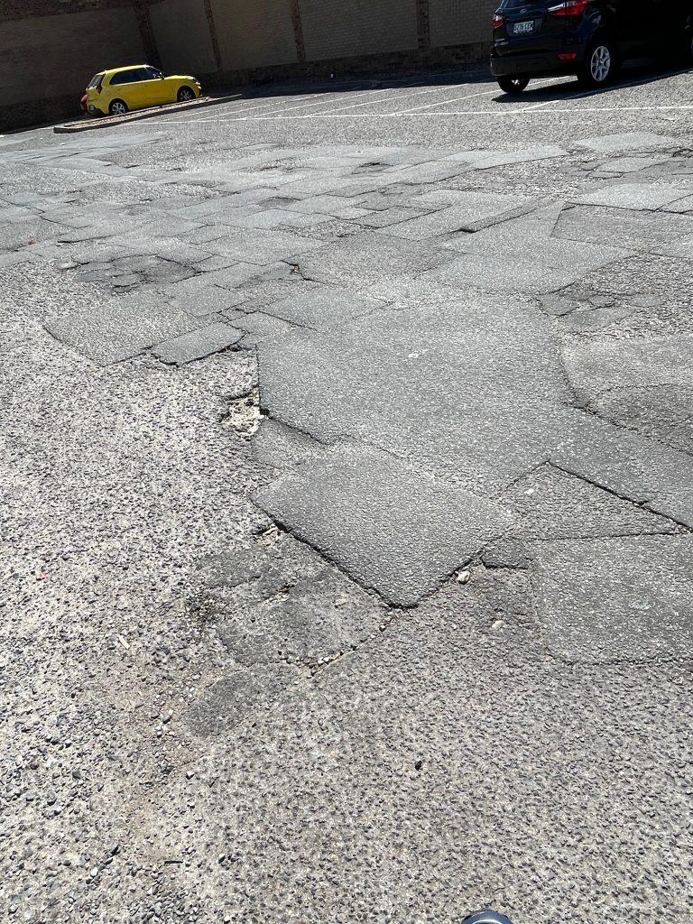 the car park is a patchwork quilt, with many patches over a long time. The background has a yellow car to the left and a black vehicle on the right. Sefton Plaza accessibility