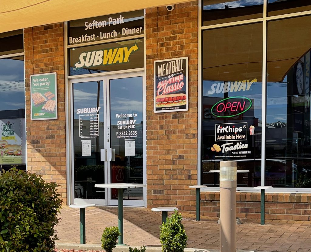 empty tables and chairs are outside subway, a double glass door on the left, and a window to the right