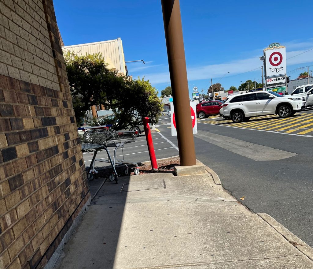left is the wall of a shop, and to the right is the carpark. Around the shop is a concrete path, and a step exit from footpath to the car park. sefton plaza accessibility