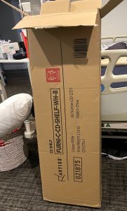 the rectangular cardboard box is being opened, and is sitting against a bed