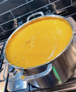 A vibrant pot of velvety orange soup bubbles atop the gas stove, its warmth radiating in the kitchen's air. The soup's rich hue contrasts against the sleek black splashback, creating an eye-catching tableau. Wisps of steam dance gracefully from the pot's surface, carrying tantalizing aromas that envelop the room. The play of light creates a mosaic of reflections on the glossy pot, adding depth to its allure. The soup's surface is adorned with subtle ripples, evidence of its gentle simmer. This vivid scene embodies both comfort and elegance, a culinary masterpiece in the making against the contemporary backdrop. Tasty Eggplant Soup.