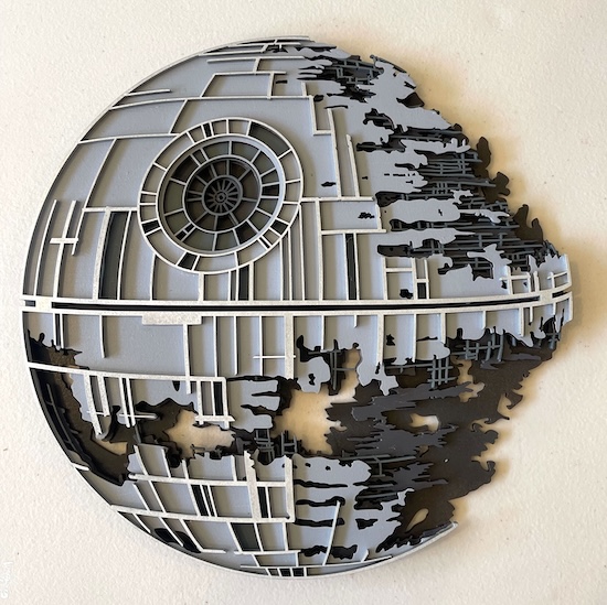 The image shows a laser-cut artwork of the Death Star from Star Wars. The circular structure is intricately cut, highlighting its iconic mechanical details, with one side appearing battle-damaged. The layered design adds depth, using grey and black elements to accentuate the details of the station’s grid-like structure. The artwork captures the distinct shape of the Death Star, making it an eye-catching piece for any Star Wars fan.