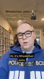 The image shows a man wearing glasses and a blue hoodie with the words “POLICE PUBLIC CALL BOX” printed across the front, referencing the TARDIS from Doctor Who. He is speaking into a small microphone clipped to his hoodie.

Text is overlaid on the image:

At the top left, it reads: “Wheelchair John Stand-up Comedy Sitting-down”, with a small cartoon figure of a man in a wheelchair next to the text.

In the middle, a caption says: “Hi, it's Wheelchair John,”

The setting appears to be indoors, with shelves full of DVDs or Blu-rays along the wall behind him, and a window letting in natural light in the background.

Disability Jokes!