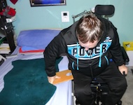 The same man leans forward from his wheelchair, placing a smooth wooden transfer board between the wheelchair and the bed. The board will help him slide safely across.