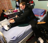 The man, still in his tracksuit, sits at the edge of the bed, stretching his leg across and using his hand to pull it closer while balancing on the transfer board. sleeping after spinal cord injury