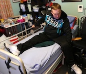 The man adjusts his body further onto the bed, one hand gripping the rail and the other resting on the mattress to steady himself. Both legs are now on the bed.