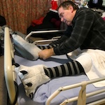 The man, now wearing a dark checkered shirt and striped socks with fluffy animal slippers, leans forward on the bed, adjusting the position of his feet and slippers. sleeping after spinal cord injury
