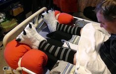 The same man lies in bed with red cylindrical cushions placed around his feet to keep them supported and separated. He still wears the striped socks and soft white animal-paw slippers. He looks down at his legs to check their position. sleeping after spinal cord injury