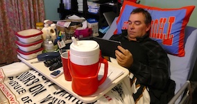 The man reclines in bed with his head propped up on a large orange pillow. A white tray table rests across his lap, holding a large red drink container, a phone, and other items. He is holding a tablet, appearing relaxed and ready to rest or read before sleep. sleeping after spinal cord injury