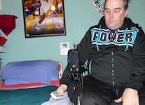 A man wearing a black tracksuit with the word “POWER” on the chest sits beside a hospital-style adjustable bed. He holds a hand control used to raise or lower the bed. The room has teal walls and a framed sports poster on the wall. sleeping after spinal cord injury